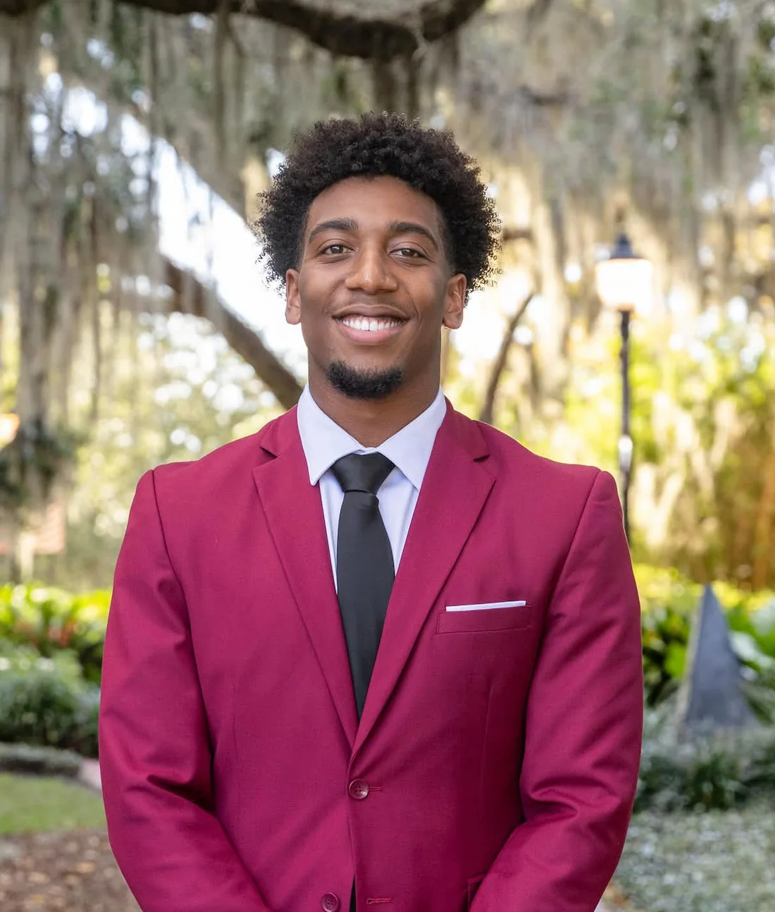 Ammiel Bowen smiling in a garnet suit and black tie, standing under oak trees draped in Spanish moss